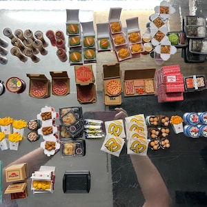 May include: A collection of miniature food items, including burgers, pizza, sushi, fries, and pretzels. There are also miniature soda cans, ramen, and other packaged foods. The items are arranged on a reflective surface.