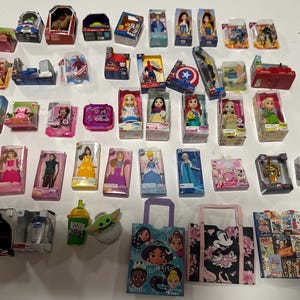 Könnte beinhalten: Eine Sammlung von Miniaturspielzeugen und Sammlerstücken, darunter Actionfiguren, Puppen und themenbezogene Waren. Die Artikel sind in Kartons verpackt und auf einer weißen Oberfläche ausgestellt. Das Sortiment umfasst Charaktere aus beliebten Franchises.