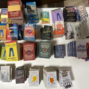 May include: A selection of miniature books, including titles such as "Sherlock Holmes," "Moby Dick," and "Little Women." The books have colourful covers and various sizes, arranged on a white surface. The collection includes classic and contemporary novels.