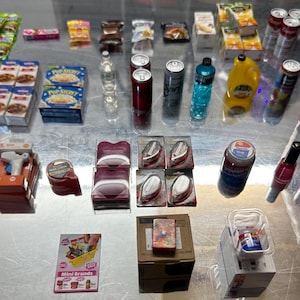 May include: A selection of miniature grocery products, including snacks, drinks, and cleaning supplies. There are small boxes of cereal, cans of fizzy drinks, bottles of water, and other packaged goods. The items are arranged on a reflective surface.