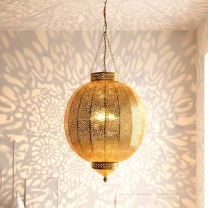 Puede incluir: Una lámpara colgante esférica dorada con intrincados patrones calados. La luz cuelga de una cadena y proyecta sombras decorativas en el techo y la pared. La lámpara tiene un remate decorativo en la parte inferior.
