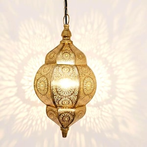 May include: A gold-coloured, ornate pendant light fitting with intricate cut-out patterns. The light casts a decorative shadow on the white background. The lamp has a bulb inside, and is suspended from a cord.