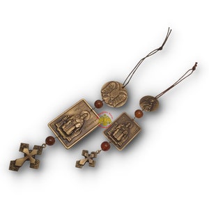 Puede incluir: Un conjunto de tres amuletos de coche dorados con iconos religiosos. Los amuletos están enhebrados con cuentas marrones y tienen un cordón marrón para colgar. Los iconos incluyen una cruz, un santo y un águila bicéfala.