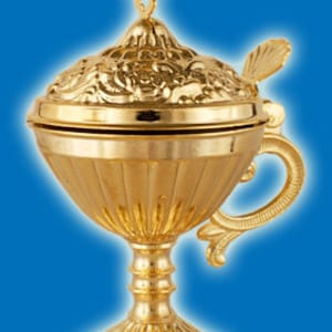 May include: A gold-colored metal censer with a decorative floral design. The censer has a lid with a cross-shaped finial and a handle on the side. The censer sits on a round base with a floral design.
