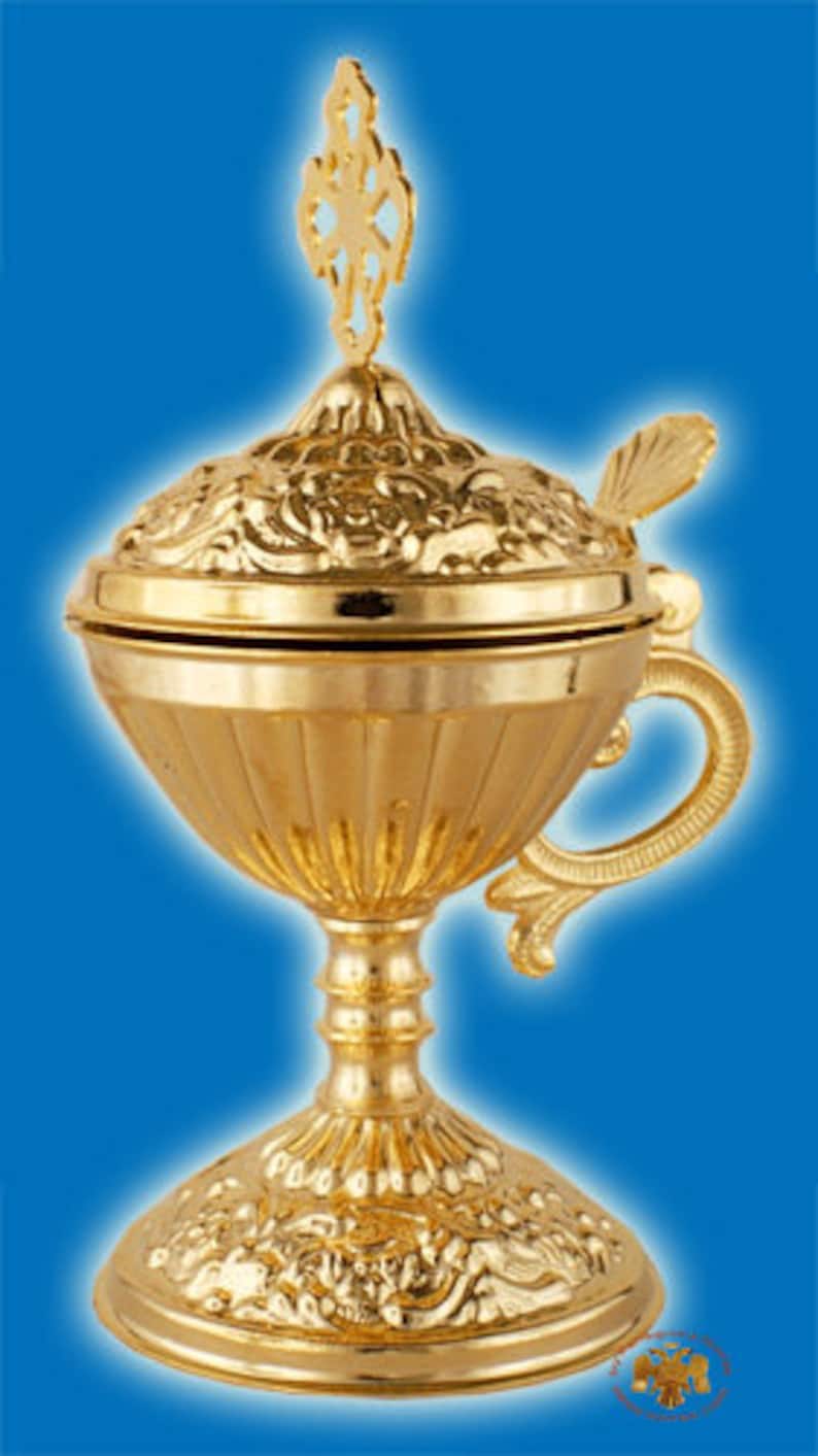 May include: A gold-colored metal censer with a decorative floral design. The censer has a lid with a cross-shaped finial and a handle on the side. The censer sits on a round base with a floral design.