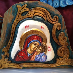 May include: A carved wooden icon depicting the Virgin Mary and baby Jesus. The icon is painted in gold, red, blue, and white. The background is a teal blue with gold leaf accents. The icon is framed in a carved wooden frame.