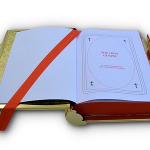 Puede incluir: Un atril de libro dorado con una cinta roja y un libro blanco abierto en una p&aacute;gina con el texto "THE HOLY GOSPEL" en rojo. El libro est&aacute; sobre un fondo blanco.