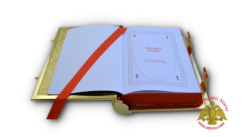 Puede incluir: Un atril de libro dorado con una cinta roja y un libro blanco abierto en una p&aacute;gina con el texto "THE HOLY GOSPEL" en rojo. El libro est&aacute; sobre un fondo blanco.
