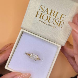 May include: A gold ring featuring a rectangular radiant-cut diamond in a bezel setting, presented inside a cream-colored jewelry box with the gold-embossed text SABLE HOUSE HEIRLOOMS printed on the interior lid lining.