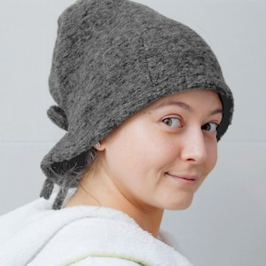 May include: A gray, felted hat with a rounded crown and a brim that extends to the sides and back. The hat has a tie detail at the back. The person is wearing a white hooded robe.
