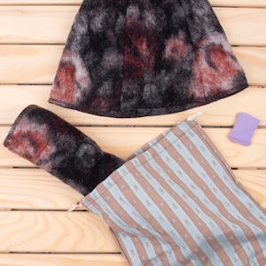 Puede incluir: Un gorro de sauna afieltrado con un patrón moteado en negro, gris y rojo, junto con una manopla de sauna enrollada a juego. También hay una bolsa con cordón a rayas y un pequeño objeto morado. Los artículos están dispuestos sobre una superficie de madera clara.