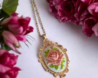 May include: A gold-coloured pendant necklace with a cameo of a pink rose on a pale green background. The pendant has an ornate, decorative frame. Pink roses are in the background.