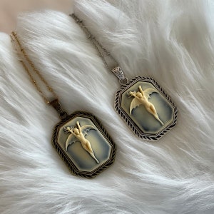 Pode incluir: Dois colares com um pingente de camafeu que representa uma figura alada. Os pingentes s&atilde;o em tom prateado e dourado com um camafeu em preto e branco.
