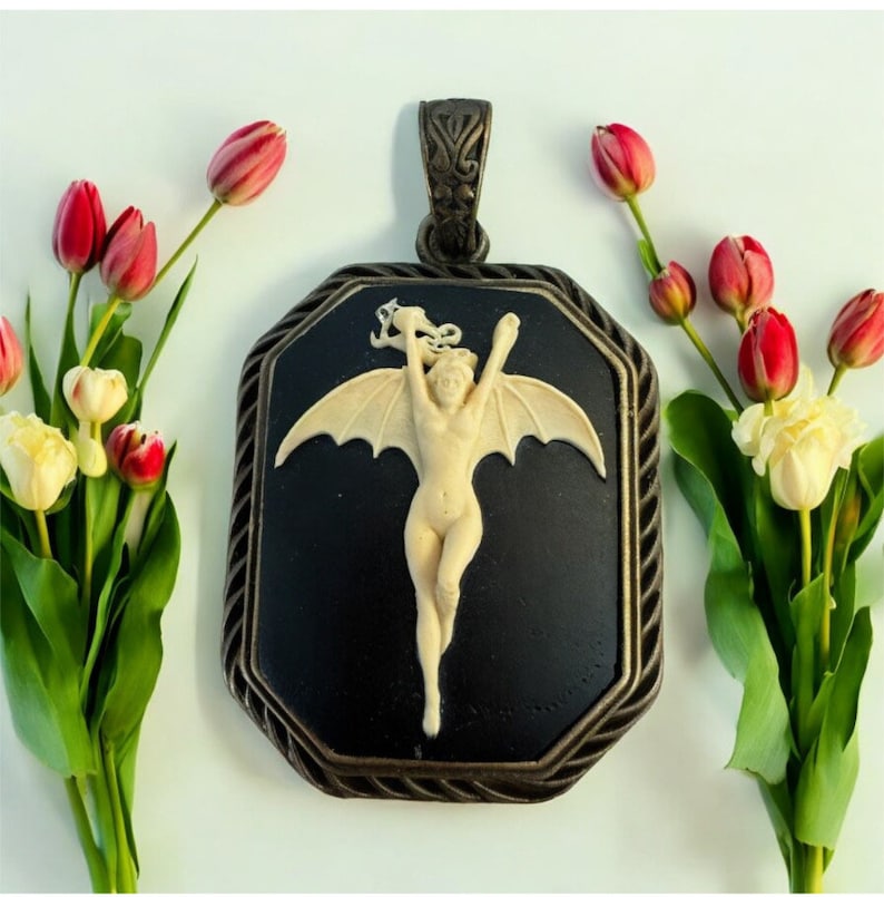 Pode incluir: Um pingente de camafeu preto com moldura prateada apresenta uma figura esculpida branca de uma mulher com asas. A mulher &eacute; representada com os bra&ccedil;os levantados e as asas abertas. O pingente &eacute; rodeado por uma borda decorativa.