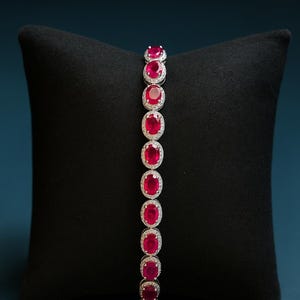 Puede incluir: Una pulsera de plata con piedras preciosas rojas de corte ovalado, cada una rodeada por un halo de piedras más pequeñas y claras. La pulsera se exhibe sobre una superficie negra, mostrando su color vibrante y su intrincado diseño.