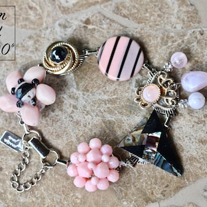 May include: A custom-sized bracelet with a variety of decorative elements. It features pink beads, a black and pink striped circle, and a black triangle with iridescent accents. The bracelet is sized from 6.5" to 9.0".