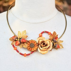 Peut inclure: Un collier en or avec un motif floral composé d'une grande rose beige, d'une petite fleur orange et de feuilles dorées. Le collier comprend également des perles rouges et orange.
