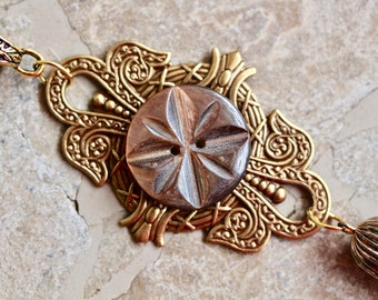 May include: A gold-toned metal pendant with an ornate design and a brown wooden button in the center. The pendant has a tassel attached to it.