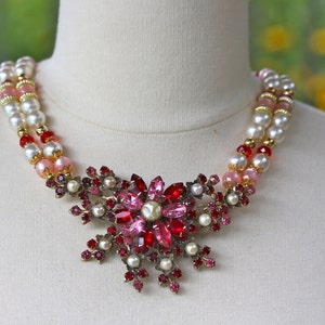 May include: A two-strand pearl necklace with pink and red beads and a large, ornate, red and pink rhinestone flower brooch.