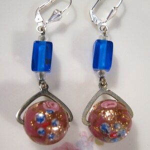 Può includere: Un paio di orecchini pendenti in argento con un cubo di vetro blu e una perla di vetro rosa con accenti dorati e blu.