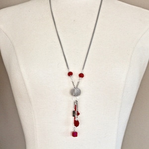 May include: A silver chain necklace with a silver charm and red beads. The charm has a textured surface and is attached to a dangling chain with more red beads.