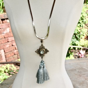 Puede incluir: Un collar de cadena plateada con un gran colgante adornado con un cristal transparente y una borla gris.