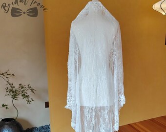 May include: A white lace bridal veil displayed on a gold stand. The veil is long, with intricate floral patterns along the edges. The Bridal Trove logo is visible in the upper left corner. The background is a warm yellow wall and wooden floor.