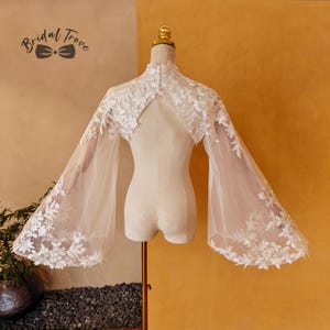 May include: A white bridal cape with bell sleeves, displayed on a mannequin. The cape features intricate floral lace detailing along the neckline and sleeve edges. The Bridal Trove logo is visible in the upper left corner.