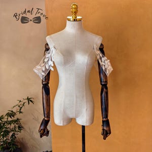 Peut inclure: Un mannequin de couture crème avec des bras en bois et un sommet doré. Le mannequin porte des manches en dentelle blanche transparente avec un motif de feuilles. Le texte "Bridal Trove" est dans le coin supérieur gauche.