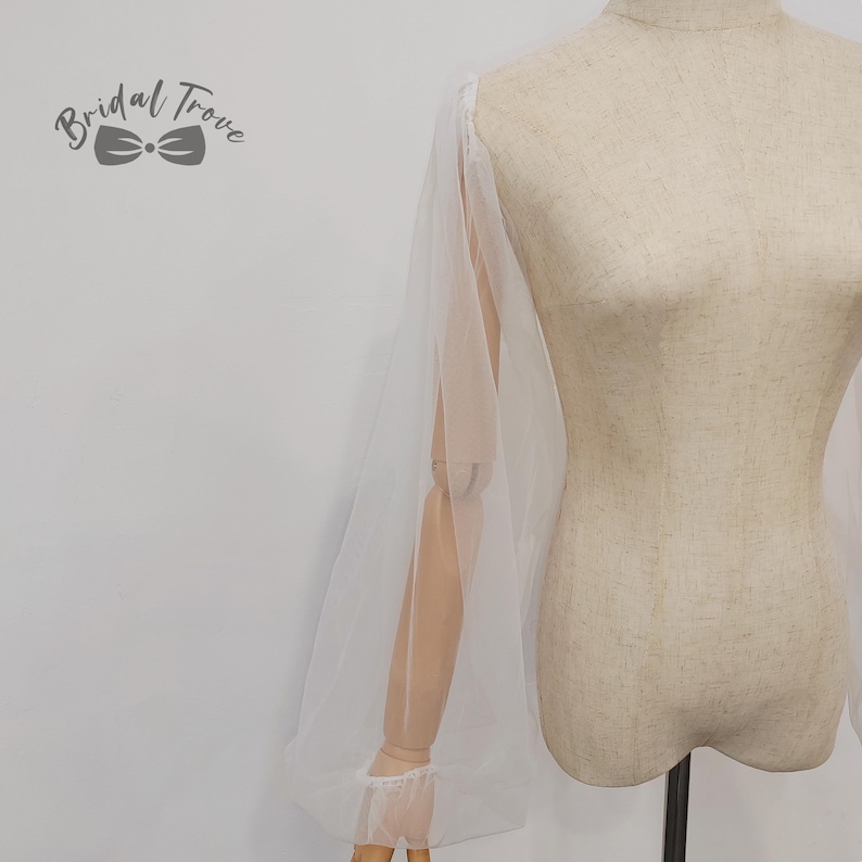 K&ouml;nnte beinhalten: Ein wei&szlig;er, durchsichtiger Braut&auml;rmel mit einem Puff-Design. Der &Auml;rmel ist an einer Schaufensterpuppe befestigt. Das Bridal Trove-Logo befindet sich in der oberen linken Ecke. Der &Auml;rmel hat eine Manschette am Handgelenk.