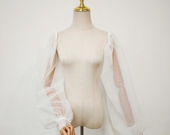 May include: A white, sheer, long-sleeved top with voluminous, puffy sleeves. The sleeves are gathered at the wrist with a button closure. The top is displayed on a mannequin with a gold-colored neck and headpiece.