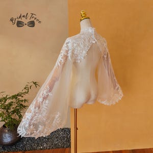 May include: A sheer, ivory bridal cape with floral lace detailing. The cape features a high neckline and wide, flowing sleeves. The cape is displayed on a mannequin against a warm, yellow background. The text "Bridal Trove" is visible in the upper left corner.
