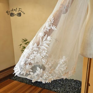 May include: A sheer, off-white bridal veil with intricate floral lace detailing along the edge. The delicate lace features a repeating pattern of flowers and leaves. The Bridal Trove logo is visible in the upper left corner.