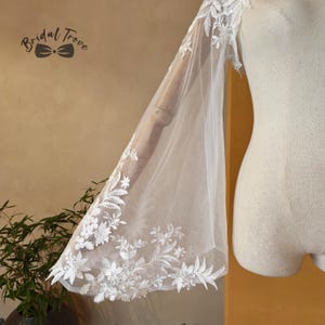May include: A white, sheer bridal sleeve with intricate floral lace detailing. The sleeve is attached to a mannequin, showcasing the delicate design and texture. The Bridal Trove logo is visible in the upper left corner.