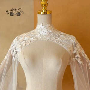May include: Ivory lace bridal cape with floral and leaf embroidery. The cape features a high neckline and sheer sleeves. The Bridal Trove logo is visible in the upper left corner. The cape is displayed on a mannequin.