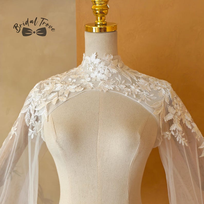 May include: Ivory lace bridal cape with floral and leaf embroidery. The cape features a high neckline and sheer sleeves. The Bridal Trove logo is visible in the upper left corner. The cape is displayed on a mannequin.