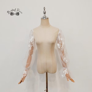 Pode incluir: Mangas de noiva brancas e transparentes com aplicações de renda floral. As mangas compridas apresentam um design bufante no ombro e um punho flare. O logotipo Bridal Trove é visível.
