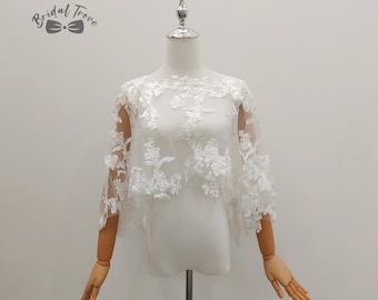 May include: A white lace bridal cape with floral embroidery is displayed on a mannequin. The cape features sheer sleeves and a delicate, intricate design. The Bridal Trove logo is visible in the upper left corner.