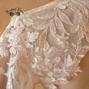 May include: Close-up of ivory lace fabric with floral and leaf embroidery. The delicate design features intricate details and a sheer mesh backing. The Bridal Trove logo is visible in the upper left corner.