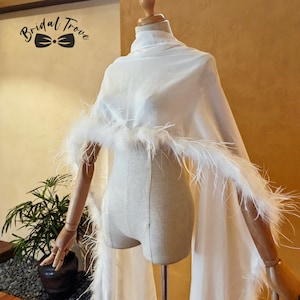 Puede incluir: Un chal blanco transparente drapeado sobre un maniquí, con cuello alto y largos paneles de tela fluida. La prenda está acentuada con un borde grueso de plumas blancas a lo largo de los extremos. El texto Bridal Trove con un icono de lazo es visible.