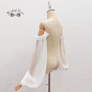 May include: A white off-the-shoulder top with long, sheer sleeves. The sleeves are gathered at the wrist, creating a voluminous effect. The top is displayed on a mannequin, showcasing its design and fit. The Bridal Trove logo is visible.