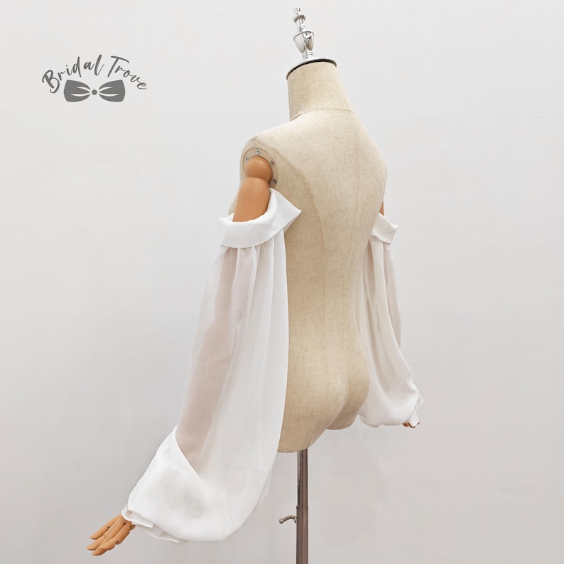 May include: A white off-the-shoulder top with long, sheer sleeves. The sleeves are gathered at the wrist, creating a voluminous effect. The top is displayed on a mannequin, showcasing its design and fit. The Bridal Trove logo is visible.
