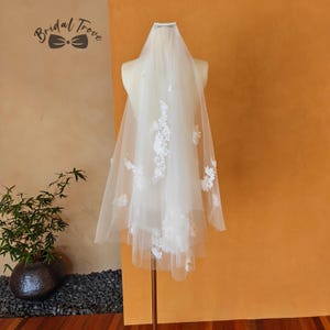 Puede incluir: Un velo de novia blanco con aplicaciones de encaje floral y perlas dispersas se exhibe en un maniquí. El velo es transparente y tiene un diseño de dos niveles. El texto "Bridal Trove" es visible en la esquina superior izquierda.