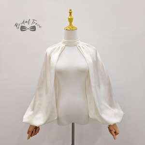 Pode incluir: Top de noiva de cetim branco com gola alta e mangas compridas e volumosas. O top é exibido em um manequim com braços de madeira. O texto "Bridal Trove" é impresso no canto superior esquerdo.