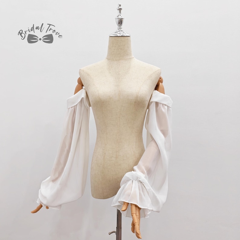 May include: White off-the-shoulder sleeves with long, flowing fabric and ruffled cuffs. The sleeves are displayed on a mannequin. The text "Bridal Trove" is in the upper left corner.