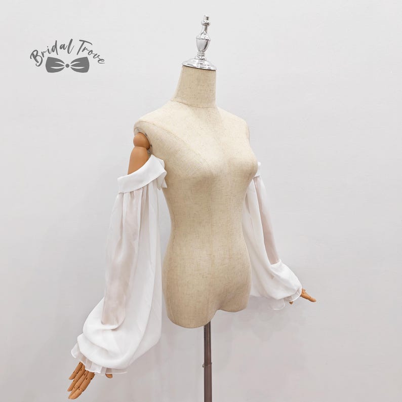 May include: A white, off-the-shoulder bridal top with long, sheer sleeves. The sleeves are gathered at the shoulders and wrists, creating a voluminous, romantic look. The top is displayed on a mannequin. The text "Bridal Trove" is in the upper left corner.