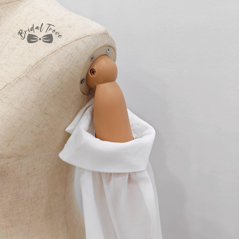 May include: A white long-sleeve shirt with a wide cuff is displayed on a mannequin. The mannequin's arm is visible, and the shirt's cuff is folded over. The background is a plain white wall. The text "Bridal Trove" is visible.
