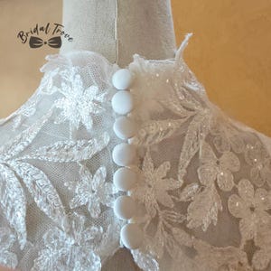 May include: Close-up of a white lace bridal top with floral embroidery and sparkling sequins. The back features a row of six round, white buttons. The Bridal Trove logo is visible in the upper left corner.