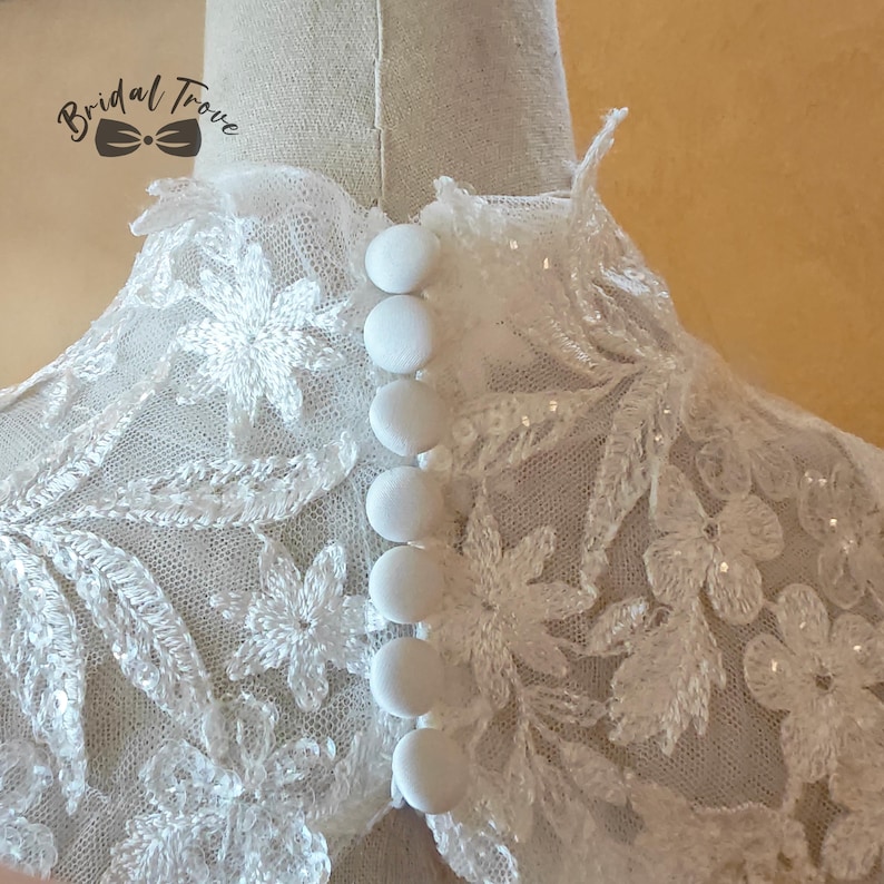 May include: Close-up of a white lace bridal top with floral embroidery and sparkling sequins. The back features a row of six round, white buttons. The Bridal Trove logo is visible in the upper left corner.