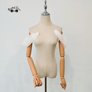 Puede incluir: Un maniquí beige con brazos y manos de madera articulados. El maniquí tiene un soporte de metal y está adornado con mangas de tela blanca. El logotipo "Bridal Trove" es visible en la esquina superior izquierda.